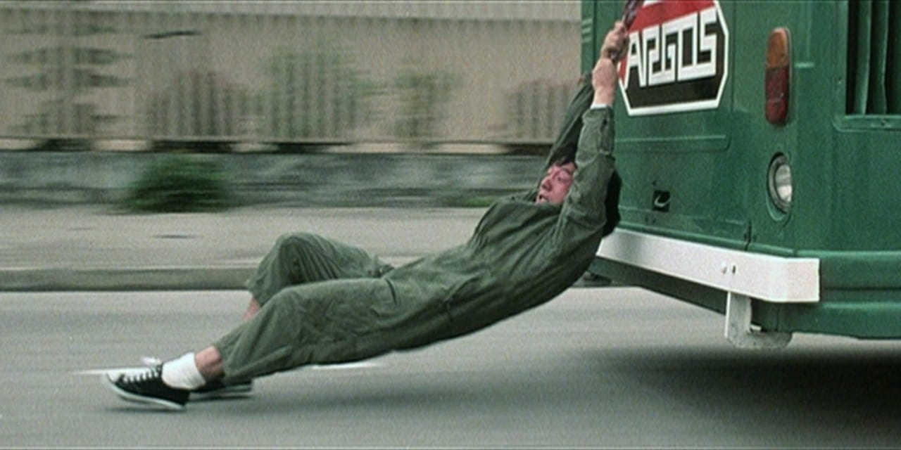 Jackie Chan hanging off of a moving bus by an umbrella, his feet dragging across the road.