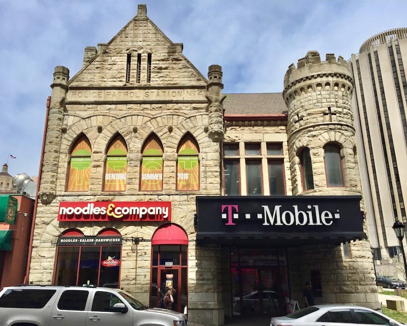 A historic stone building with a turret on the right side. Engraved in the stone is Police Patrol Station No. 4. The former theater's marquee is now a T Mobile sign, and another sign for Noodles & Company hangs on the stone face of the building.