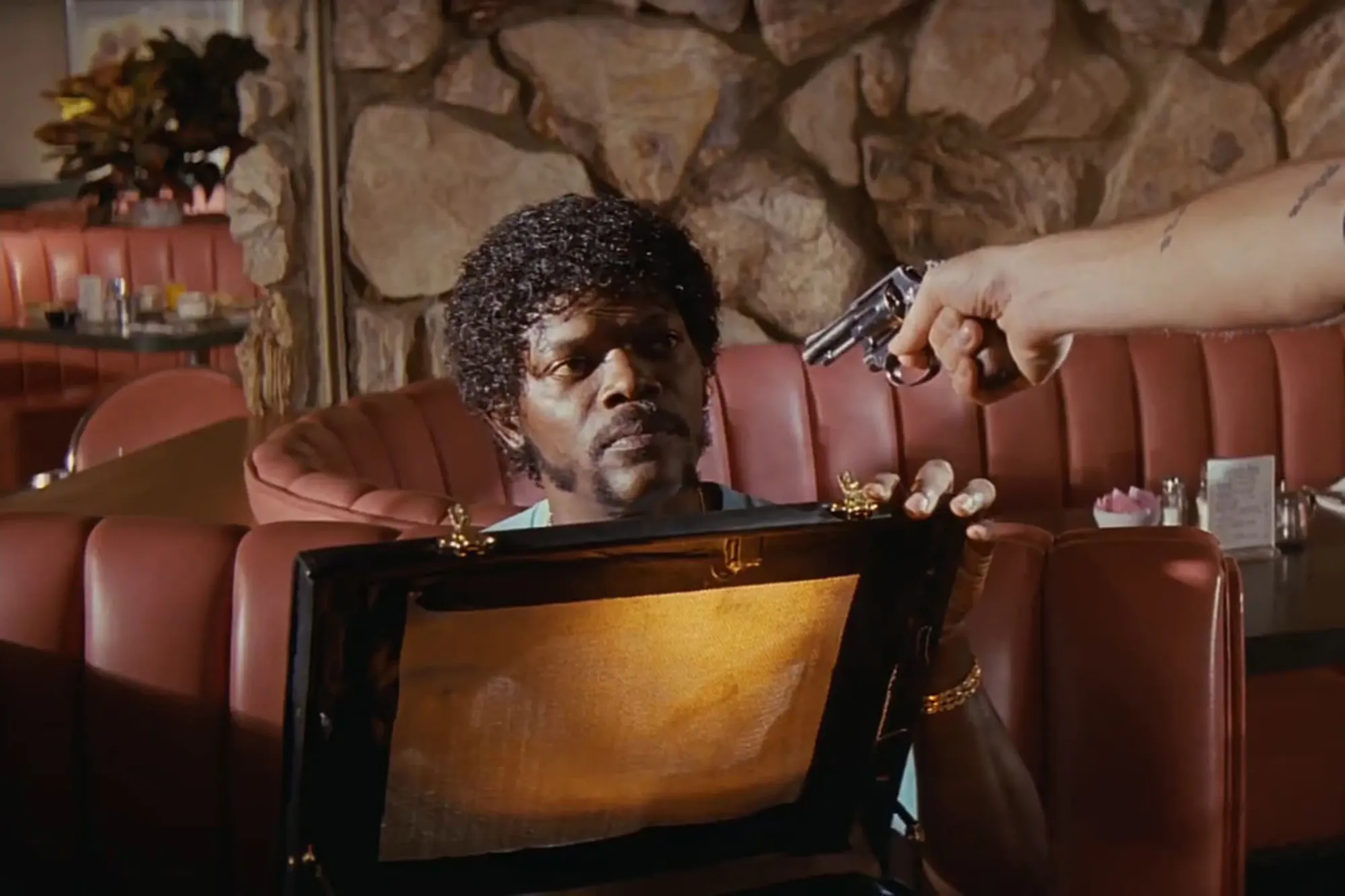 A shot from Pulp Fiction. Jules sits at a diner booth, holding open a briefcase that glows with golden light. A gun is pointed at his head by an arm emerging from offscreen.