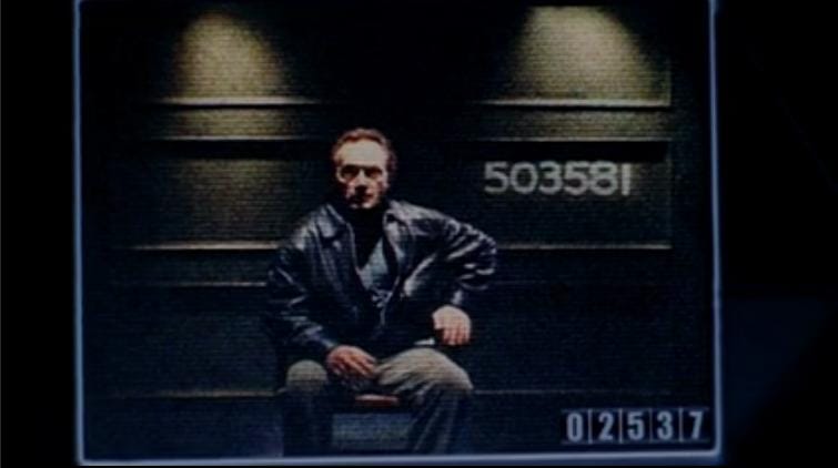 A grainy image of a man in a chair, with a number (503581) on the wall behind him. On the bottom of the screen, another number (02537)