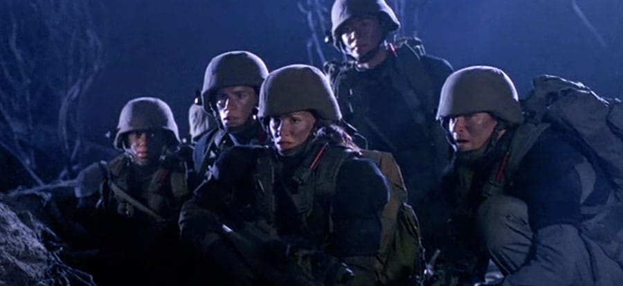 The cast of Space Above and Beyond on an alien planet, in military uniforms and helmets. They are crouched and looking at something offscreen.