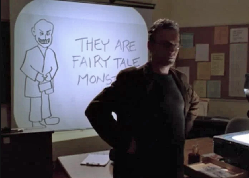 Giles, standing in a classroom, with an overhead projector shining on the screen behind him. The image is a creepy man and the words "They are fairy tale monsters".