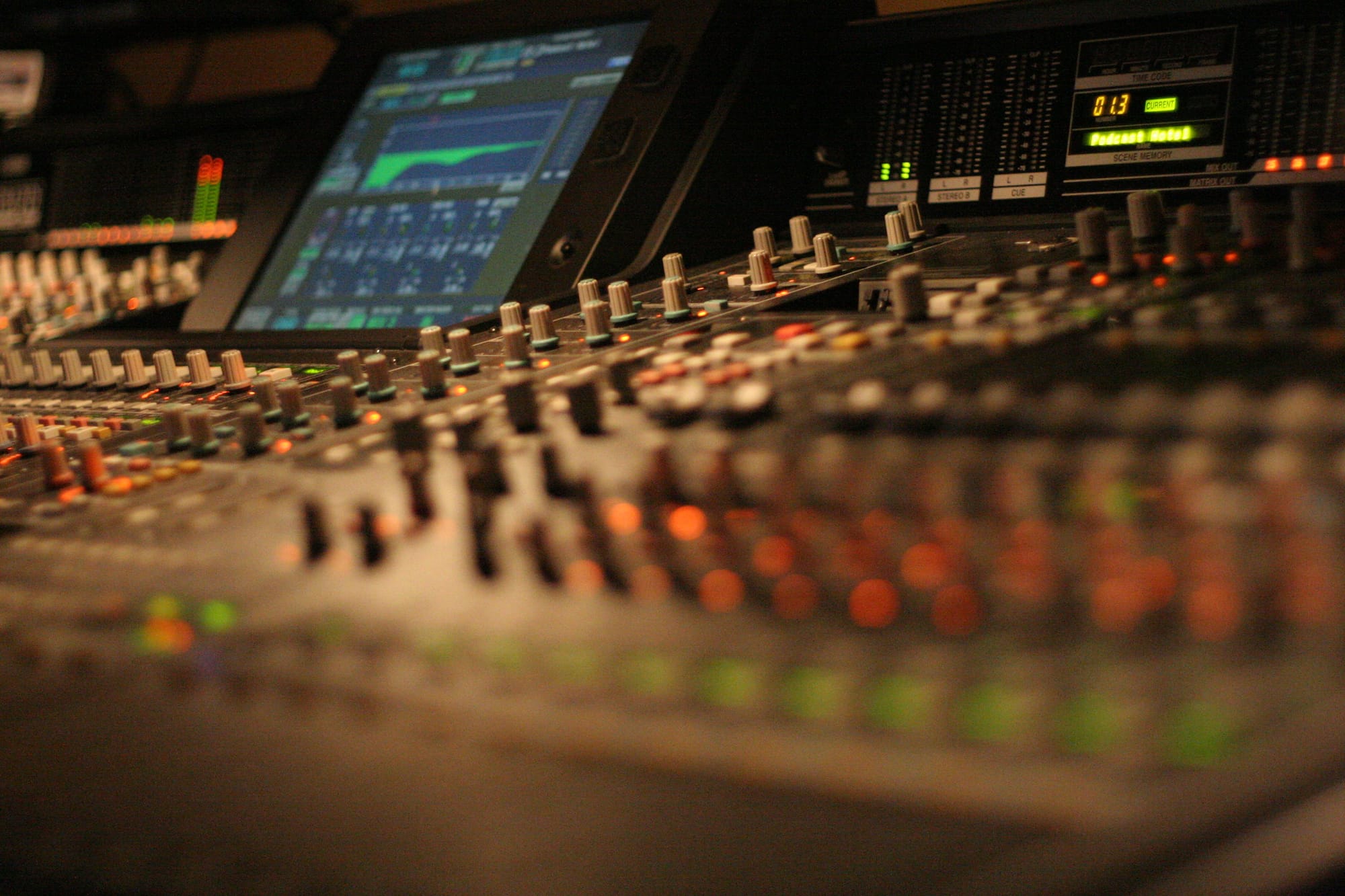 A close up of an audio mixing board, the foreground out of focus, and becoming in focus the further it goes back where we can see a monitor.