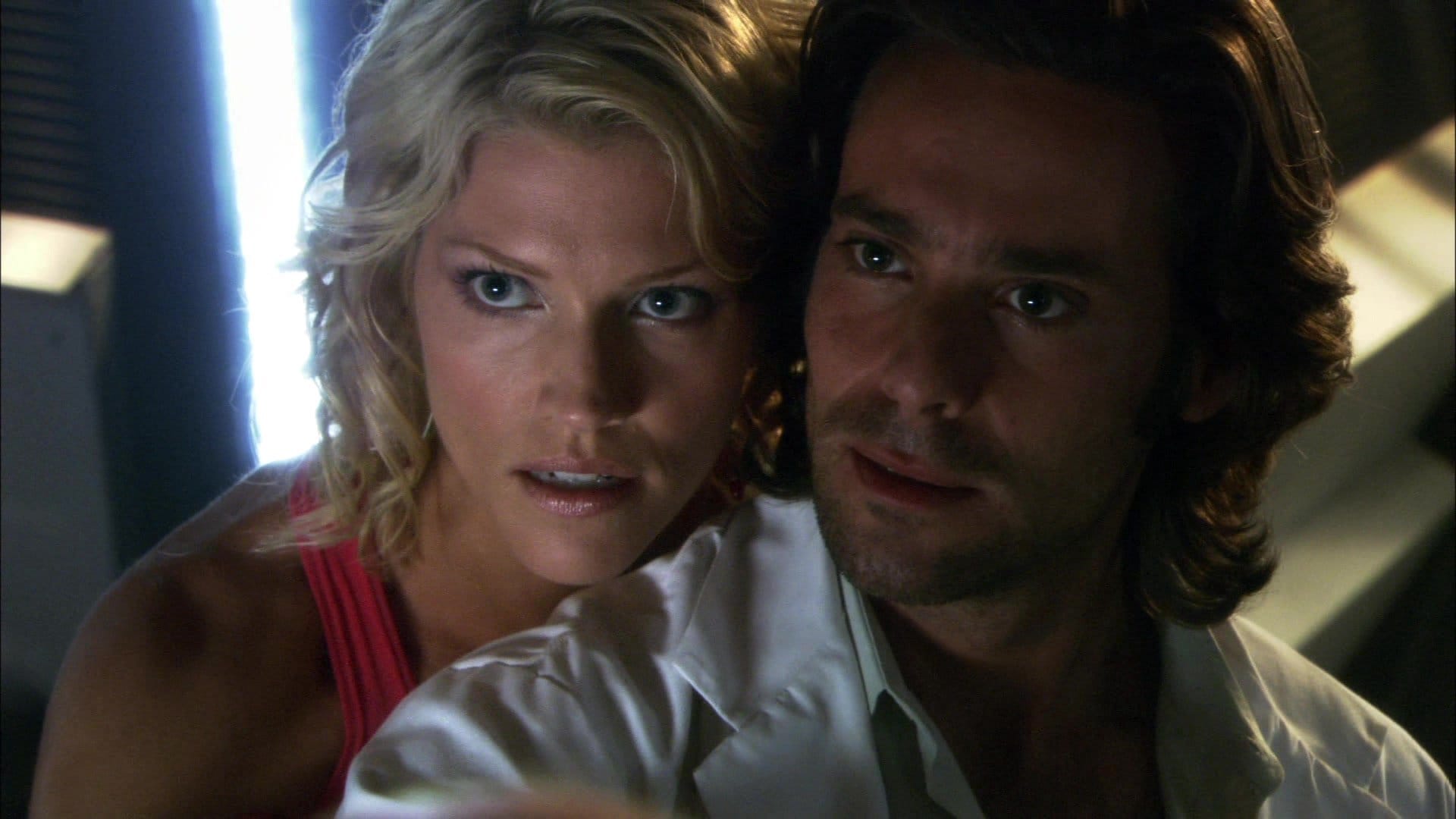 A screencap of Baltar with Head Six in her red dress leaning over his shoulder, a closeup of both of their heads.