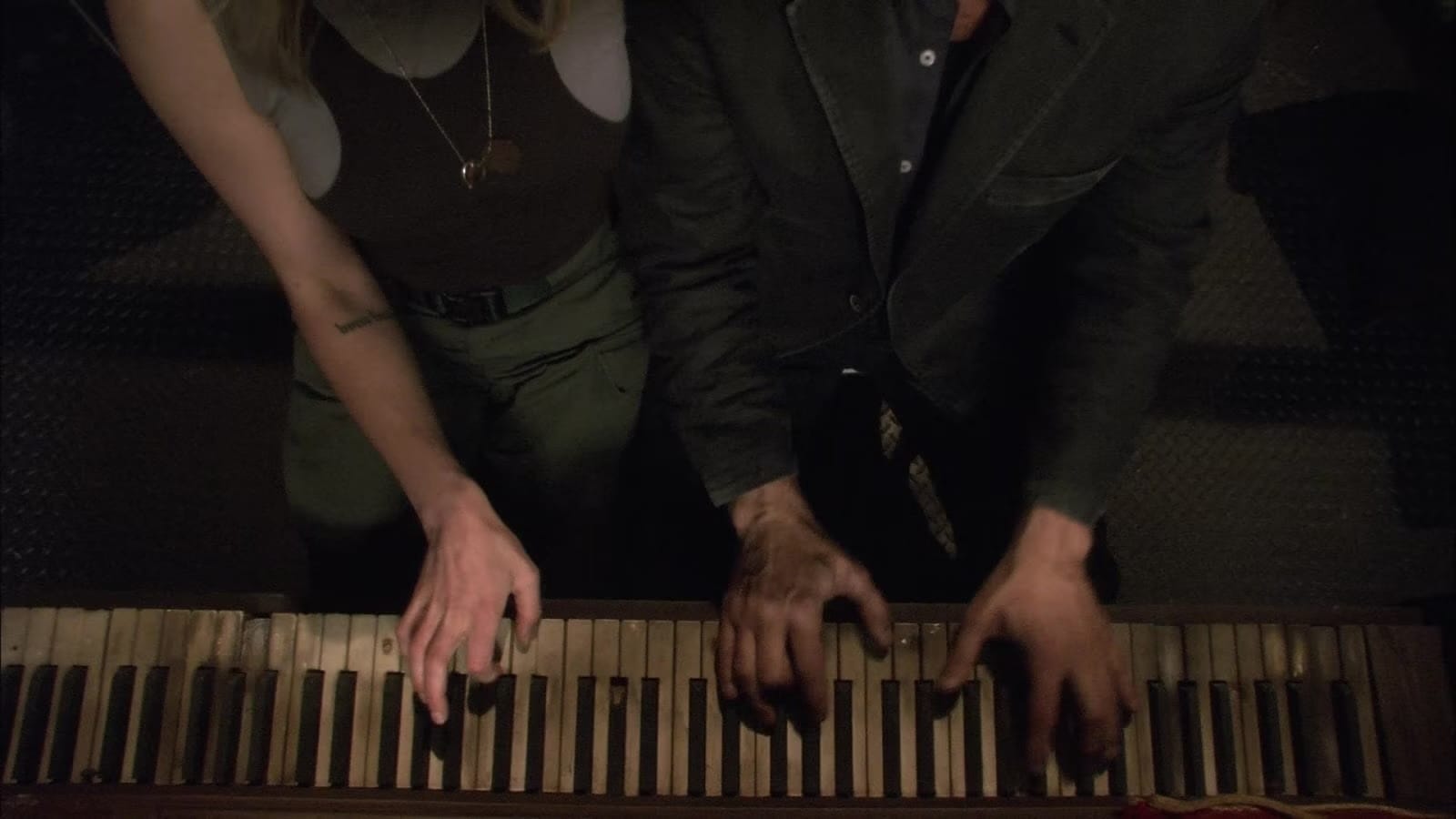 An overhead shot of two people at a beaten up piano, only their arms and hands on the keyboard are visible