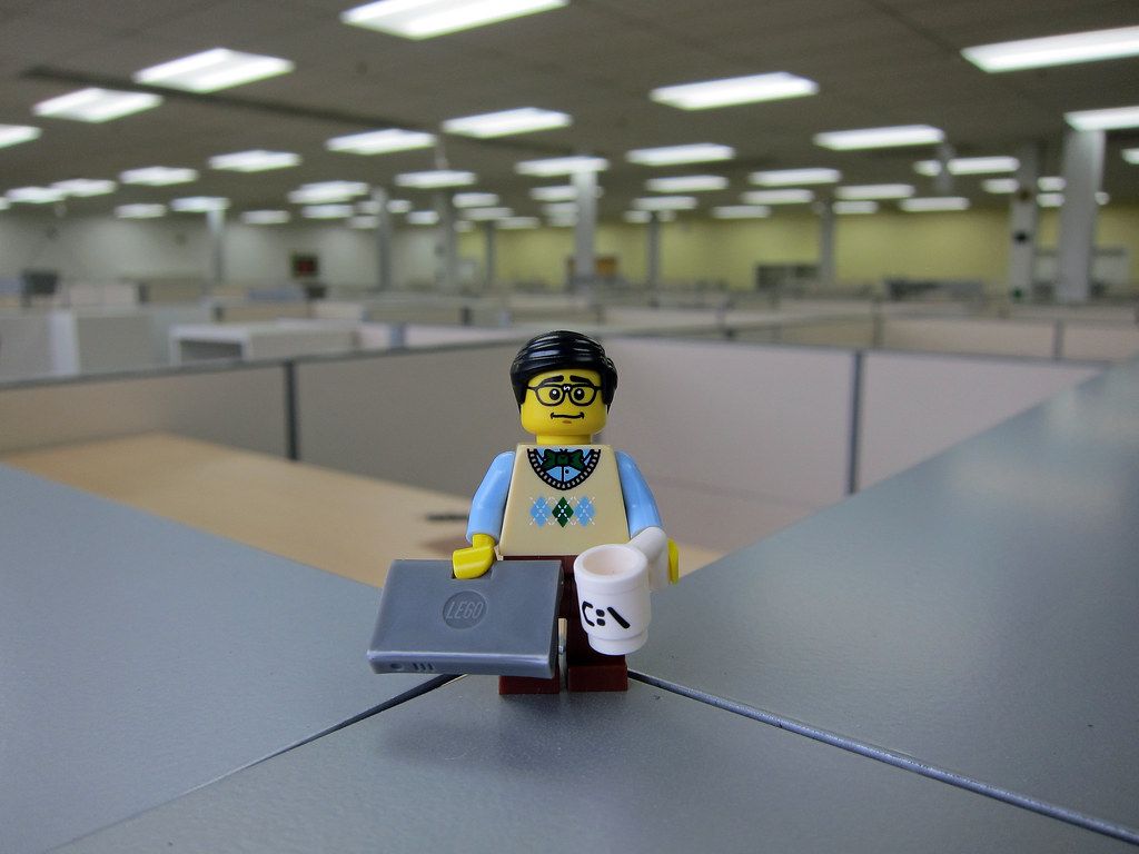 A lego figurine of a programmer with a laptop and coffee mug, standing in an office.