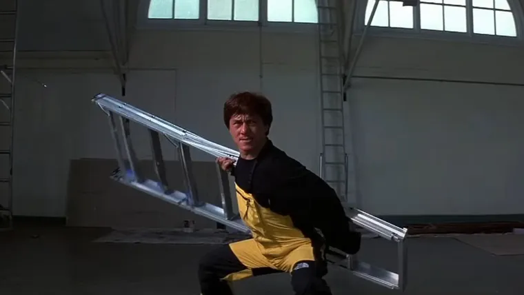 Jackie Chan in First Strike. He's wearing a yellow and black jumpsuit and holding a folded-up stepladder across his back in a combat pose, the way someone would hold a staff.