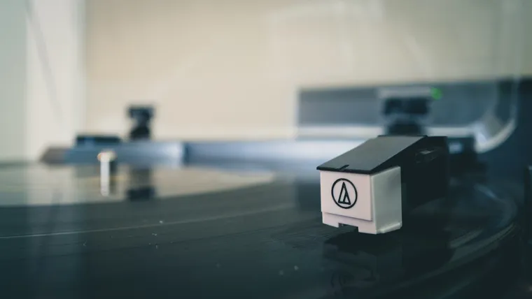 A close up of a vinyl record on a record player, largely out of focus, with the needle head in focus in the right foreground.